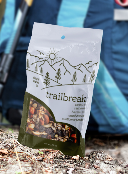 Hand holding a Trailbreak trail mix pouch outdoors with mountain and pine tree artwork